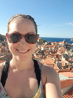 Selfie of a woman in sunglasses smiling in front of a hilly seaside town filled with white buildings and terracotta roof tiles.